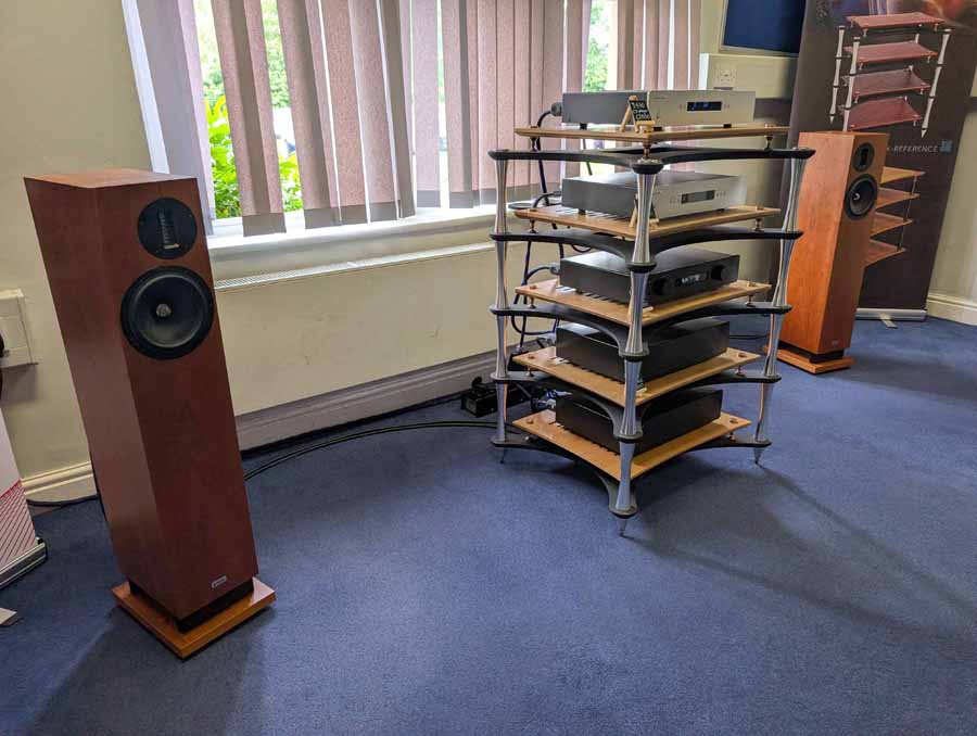 HiFi NETWORK
Victor, from HiFi Network, talked me through this cool, ultra-simple system. The stars of the show were a pair of ELAC Debut Reference 3.0 (£1399), which they claim, “builds on the success of Debut 3.0 with redesigned driver technology, refined cabinet design, and a true 3-way floorstanding architecture.” Also in the range, both with 2-way bass reflex, are: standmounts (£749) and a centre channel speaker (£449). These come in five different finishes, including: black, green, satin white, titanium, and walnut.
An AVM CS30.3 streaming amplifier was doing an admirable job of driving the ELACs, even with some laid back, and gentle smooth jazz. Having reviewed the Carina BS 243.4 bookshelf loudspeakers from the brand, I can testify their speakers benefit from an amp that can drive them, to bring out their best. This amp had it covered, with ease, and was also a lovely-looking box in chrome finish (£3700). Also on display, but not in the system at the time I was in the room, was an Inspiration C2.3 streaming amp, in a gorgeous gold satin colour. The option of this finish is exclusive to this model but doesn’t carry any additional premium. Gorgeous kit.